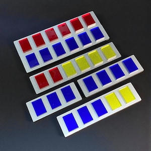 Könnte beinhalten: Vier Sätze rechteckiger, silberfarbener Balken mit farbigen rechteckigen Einsätzen. Die Einsätze sind rot, blau und gelb. Die Balken sind auf einer dunklen Oberfläche angeordnet.