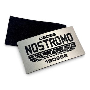Peut inclure: Une plaque rectangulaire en métal argenté avec une gravure noire indiquant USCSS NOSTROMO et 180286, accompagnée d'un emblème ailé noir. La plaque est placée partiellement sur une pièce de tissu rectangulaire noire à l'aspect feutré.