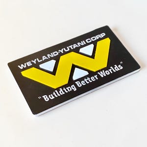 May include: A rectangular black patch featuring the yellow Weyland-Yutani Corp logo and the white text "Building Better Worlds" with two small white triangular accents positioned above the yellow W-shaped graphic element.