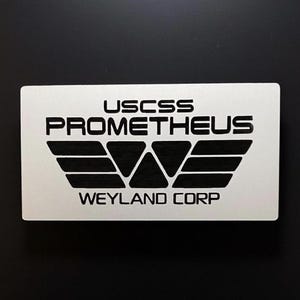May include: A rectangular silver-toned plaque featuring black engraved text that reads USCSS PROMETHEUS and WEYLAND CORP, separated by a stylized black wing-shaped logo consisting of five triangular segments arranged in a horizontal formation.