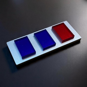 May include: A silver-coloured rectangular object featuring three coloured rectangular blocks. Two blue blocks and one red block are arranged on the silver base. The blocks are evenly spaced and have a glossy finish.