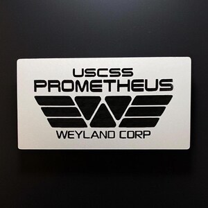 May include: A white rectangular sign with black text and a black logo. The text reads "USCSS PROMETHEUS WEYLAND CORP". The logo is a stylised wing with a central triangle.