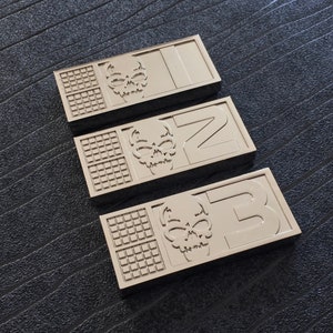 Może przedstawiać: Trzy prostokątne srebrne metalowe tabliczki z motywem czaszki i literą. Tabliczki są ułożone w stos, przy czym górna tabliczka pokazuje literę "N", środkowa tabliczka pokazuje literę "Z", a dolna tabliczka pokazuje literę "B".