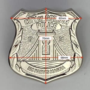 May include: A silver-coloured metal police badge with the text "CITY OF GOTAM POLICE" and "SIC PARVIS MAGNA". The badge features a stylised image of a bridge with the numbers "17" and "24" on either side. The badge measures 62 mm wide and 70 mm tall.