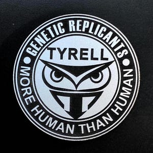May include: A circular silver sticker featuring the text "GENETIC REPLICANTS" and "MORE HUMAN THAN HUMAN" around an owl design. The word "TYRELL" is above the owl graphic.