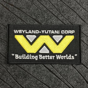 Puede incluir: Parche rectangular negro con el texto "WEYLAND-YUTANI CORP" sobre un logotipo amarillo y plateado. Debajo del logotipo, el texto dice "Building Better Worlds". El parche está sobre una superficie negra texturizada.