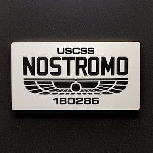 May include: A rectangular silver-toned metal plaque featuring black engraved text and graphics. The top line reads USCSS, the center displays the word NOSTROMO above a stylized winged emblem, and the bottom line features the number 180286.