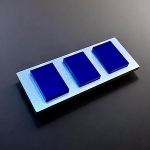 Może przedstawiać: Prostokątny, srebrny przedmiot z trzema żywymi, niebieskimi prostokątnymi blokami ułożonymi w rzędzie. Bloki są równomiernie rozmieszczone i wydają się być wykonane z półprzezroczystego materiału. Tło jest ciemnoszare.