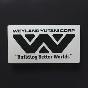 May include: A rectangular silver-colored plaque featuring the black printed text WEYLAND-YUTANI CORP at the top, a large stylized black letter W logo in the center, and the black text "Building Better Worlds" at the bottom of the surface.