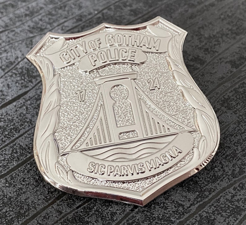 May include: A silver metal police badge with the words "City of Gotham Police" and "Sic Parvis Magna" inscribed on it. The badge features a stylised bridge with the numbers "17" and "24" on either side.