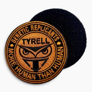 May include: A circular embroidered patch featuring a gold and black owl emblem with the text TYRELL, GENETIC REPLICANTS, and MORE HUMAN THAN HUMAN. The patch has a black hook-and-loop backing visible behind the main circular embroidered design.