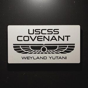Può includere: Una targa rettangolare in metallo color argento con testo inciso in nero che recita USCSS COVENANT sopra un emblema alato stilizzato e il testo WEYLAND YUTANI. Il design è pulito e industriale con lettere e grafiche nitide e chiare.