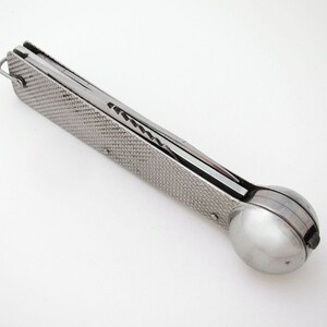 May include: A silver-toned metal kitchen tool featuring a long, rectangular handle with a textured, cross-hatched pattern and a small metal loop at one end. The opposite end features a smooth, hemispherical scoop attached to the handle.
