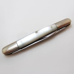 May include: A closed folding pocket knife featuring a smooth, iridescent white mother-of-pearl handle inlay secured by three small metal pins. The handle is capped with polished silver-toned metal bolsters at both ends, creating a sleek, elongated profile.