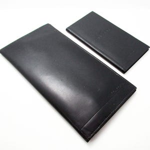 Puede incluir: Dos carteras de cuero negro. La cartera más grande es rectangular y larga, con una superficie lisa. La cartera más pequeña también es rectangular. Ambas carteras parecen estar hechas de cuero de alta calidad.