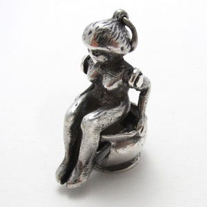 May include: A small, silver-toned charm depicting a seated figure. The figure is wearing a hat and is seated on a small, round object. The charm has a loop at the top for attachment to a necklace or bracelet. The charm is made of a shiny metal.