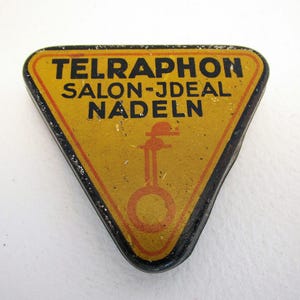 Puede incluir: Un contenedor metálico triangular vintage con el texto "TELRAPHON SALON-JDEAL NADELN" en letras negras sobre fondo amarillo. El contenedor tiene un borde rojo y un borde negro. Una ilustración naranja de un instrumento musical está en el centro.