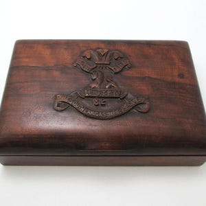 Może przedstawiać: Prostokątne drewniane pudełko o ciemnobrązowym wykończeniu. Wieko zdobi rzeźbiony emblemat z napisami "EGYPT" i "THE SOUTH LANCASHIRE REGIMENT 82". Pudełko ma gładki, wypolerowany wygląd.