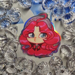 Può includere: Un adesivo olografico raffigurante un personaggio dei cartoni animati con capelli rossi e occhi blu, che fa un bacio. L'adesivo ha un contorno argentato ed è circondato da cristalli trasparenti e sfaccettati.