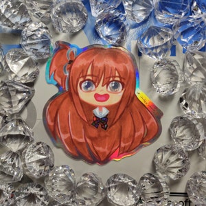 May include: A holographic sticker featuring a cartoon illustration of a girl with long brown hair, wearing a white shirt with a colourful bow tie. The girl has a big smile and is looking at the viewer.