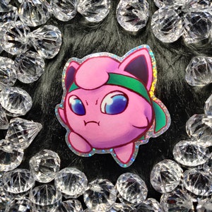 May include: A holographic sticker of a pink and green cartoon Pokemon character with a green headband. The character has a grumpy expression and is looking to the left.