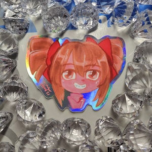 May include: A holographic sticker featuring a cartoon illustration of a girl with red hair, wearing a red bow and a pink shirt. The girl has a big smile and is looking at the viewer.
