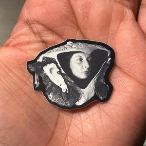 May include: Black and white sticker featuring a portrait of a woman with a hat. The image is a close-up, showing the woman's face and part of her shoulders. The sticker has a black border and is held in a hand.