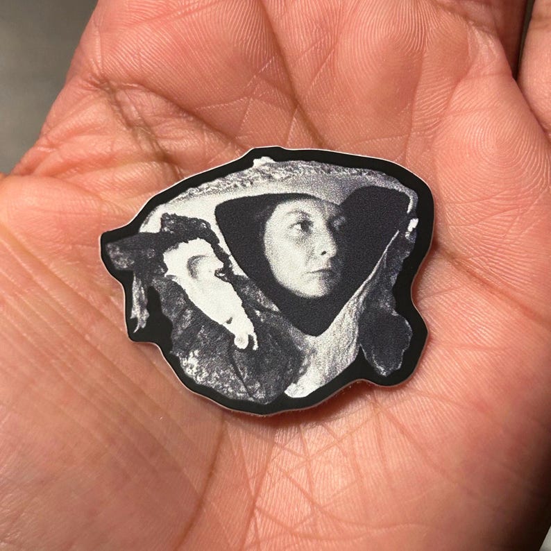 May include: Black and white sticker featuring a portrait of a woman with a hat. The image is a close-up, showing the woman's face and part of her shoulders. The sticker has a black border and is held in a hand.