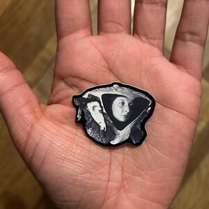 May include: A black and white sticker featuring a portrait of a woman wearing a hat. The sticker has a unique shape and is held in a person's hand. The image is a close-up shot, highlighting the details of the sticker.
