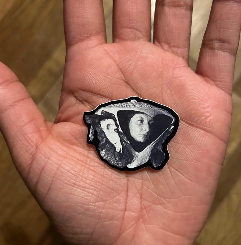 May include: A black and white sticker featuring a portrait of a woman wearing a hat. The sticker has a unique shape and is held in a person's hand. The image is a close-up shot, highlighting the details of the sticker.
