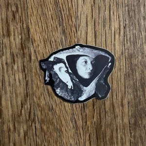 May include: Black and white sticker featuring a portrait of a woman wearing a hat. The sticker has a black border and is placed on a wooden surface. The woman's face is the focal point, with detailed shading.