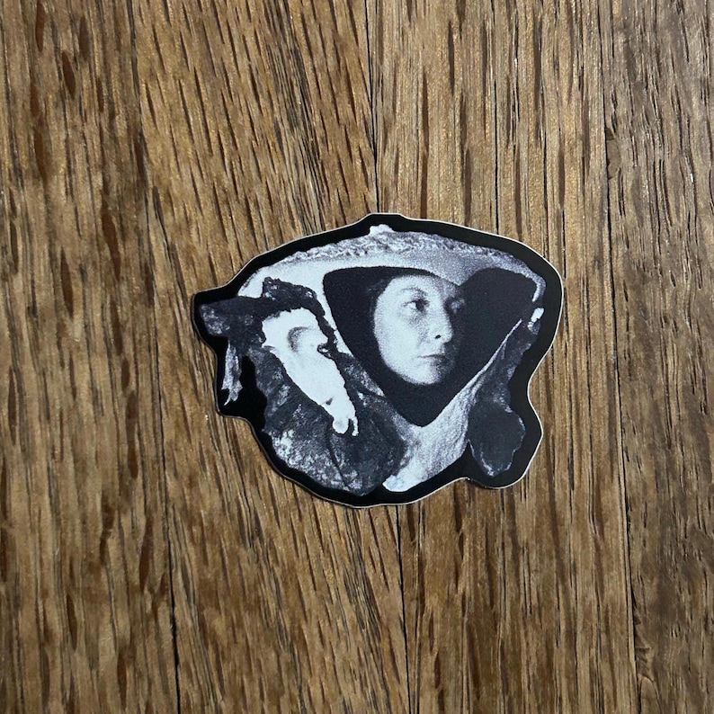May include: Black and white sticker featuring a portrait of a woman wearing a hat. The sticker has a black border and is placed on a wooden surface. The woman's face is the focal point, with detailed shading.