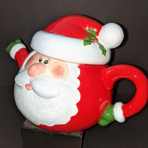 May include: Red and white ceramic teapot shaped like Santa Claus with a green holly sprig on his hat. The teapot has a white beard and a green handle.