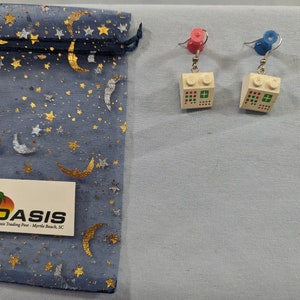 May include: A pair of white Lego brick earrings with red, green, and white details. The earrings are hanging from silver hooks with pink and blue beads at the top.