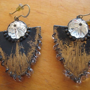 May include: A pair of black and gold earrings with a clear crystal center and a fringe of beads. The earrings are shaped like a triangle with a pointed bottom.