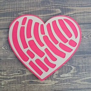 K&ouml;nnte beinhalten: Ein herzf&ouml;rmiger Papierauschnitt in leuchtendem Pink, mit einem einzigartigen Muster aus gebogenen Linien und Formen. Der Ausschnitt ist auf einer hellbraunen R&uuml;ckseite angeordnet, was einen Kontrast erzeugt. Der Hintergrund ist eine Holzoberfl&auml;che.