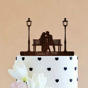 May include: A wooden wedding cake topper featuring a couple kissing on a bench under two streetlights. The topper includes the text "October 10, 2020".