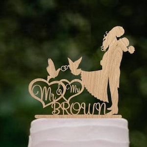 K&ouml;nnte beinhalten: Ein h&ouml;lzerner Hochzeitstortenaufsatz mit einer Silhouette eines Brautpaares, das sich die H&auml;nde h&auml;lt, zwei V&ouml;geln und dem Text "Mr. & Mrs. Brown".