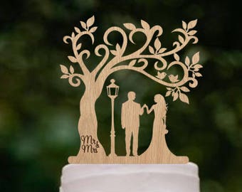 Puede incluir: Un pastel de boda de tres niveles de color morado degradado con un adorno de pastel de madera que representa a una pareja de pie bajo un árbol con un pájaro posado en una rama. El adorno dice "Mr & Mrs".