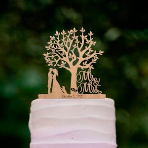 May include: A three-tier ombre wedding cake with purple frosting. The cake is decorated with a wooden topper featuring a couple standing under a tree with the words "Mr. & Mrs." and the date "October 1, 2022".