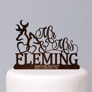 May include: A white two-tiered wedding cake with a wooden cake topper featuring a deer silhouette, the words "Mr & Mrs Fleming" and the date "25/08/2017". The cake is decorated with white flowers and berries.