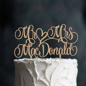 May include: A two-tiered white wedding cake with a rustic buttercream frosting. The cake is decorated with sprigs of rosemary and sugared cranberries. The cake topper says "Mr & Mrs MacDonald" in cursive.
