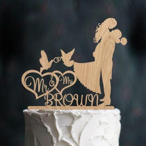 K&ouml;nnte beinhalten: Holz-Tortenaufsatz f&uuml;r Hochzeit mit einem sich umarmenden Paar, zwei Tauben mit einem Ring und dem Text "Mr. & Mrs. Brown".