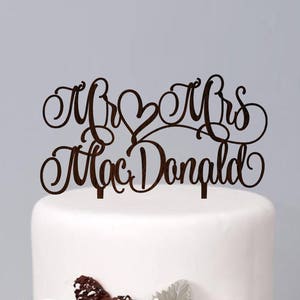 May include: A two-tiered white wedding cake decorated with white flowers, pine cones, and berries. The cake topper is a wooden sign that reads "Mr & Mrs MacDonald".