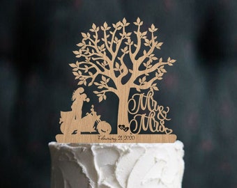 Puede incluir: Un pastel de boda blanco de dos niveles con un adorno de pastel de madera que presenta una pareja montando una motocicleta debajo de un árbol con las palabras "Mr. & Mrs." y la fecha "February 21, 2020". El pastel está decorado con ramitas de romero y bayas rojas.