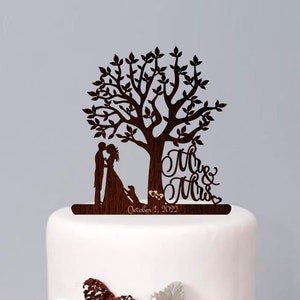 Op de afbeelding: Een witte bruidstaart met twee lagen met een houten boomtopper met een stel, een hond en de tekst "Mr. & Mrs. October 1, 2022". De taart is versierd met witte bloemen, dennenappels en bessen.