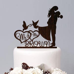 K&ouml;nnte beinhalten: Ein h&ouml;lzerner Hochzeitstortenaufsatz mit einer Silhouette eines sich umarmenden Paares, zwei V&ouml;geln mit einem Ring und dem Text "Mr. & Mrs. Brown".