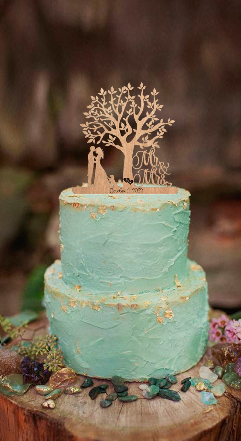 May include: A wooden wedding cake topper featuring a couple standing under a tree with a dog at their feet. The topper says "Mr. & Mrs." and "October 1, 2022".