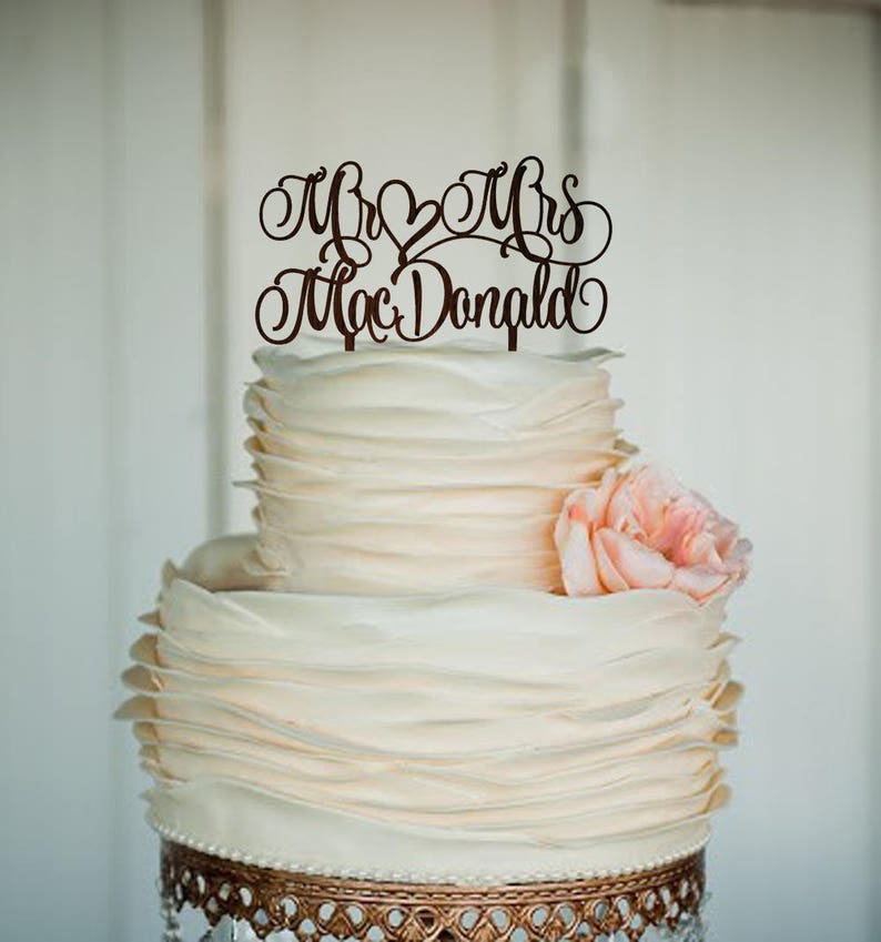 May include: A white wedding cake with a ruffled frosting design. The cake is decorated with a brown wooden topper that reads "Mr & Mrs Mac Donald".