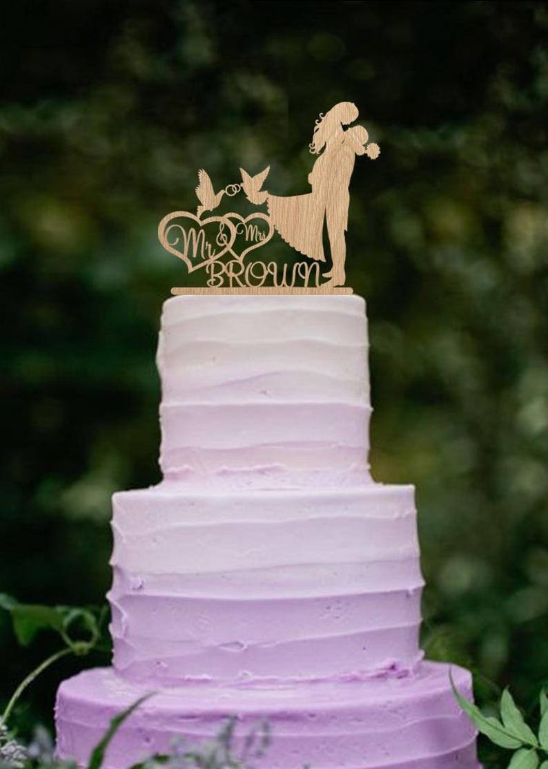 K&ouml;nnte beinhalten: Ein h&ouml;lzerner Hochzeitstortenaufsatz mit einer Silhouette eines Brautpaares, das sich die H&auml;nde h&auml;lt, zwei V&ouml;geln und dem Text "Mr. & Mrs. Brown".